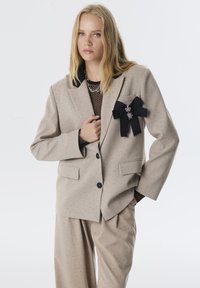 Beige pak met een gestructureerd patroon, zwarte revers, een bogenaccent op de borstzak, een dubbelgebreid ontwerp en een slouchy pasvorm van de broek.