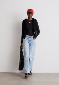 Gilet nero senza maniche sopra un top nero aderente, jeans azzurri, cappellino rosso e scarpe con stampa leopardata. Tiene una borsa tote nera. Sfondo minimalista.