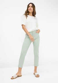 Hellgrüne, enganliegende Jeans in cropped Länge, kombiniert mit einem weißen Kurzarmoberteil mit feinem Muster und flachen weißen Sandalen.
