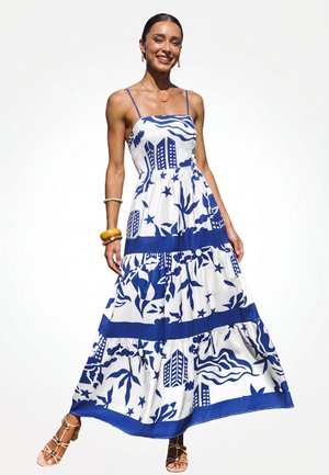 Frau lächelt, trägt ein weißes Maxikleid mit blauen abstrakten Stadtlandschafts- und Pflanzenmustern, braune Sandalen und goldene sowie hölzerne Armbänder.