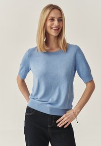 Pulover tricotat cu mânecă scurtă de culoare albastru deschis, cu decolteu rotund, manșete și tiv cu zimți. Materialul pare moale, cu o textură netedă. Se poartă blugi negri.