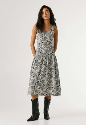 Femme portant une robe midi sans manches à imprimé floral avec jupe froncée et bottines noires à talons mi-mollet, debout devant un fond uni.