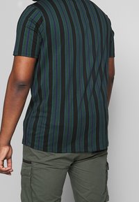 Hombre con una camiseta negra con rayas verticales verdes y azules y pantalones cargo color verde olivo, de pie contra un fondo gris liso.
