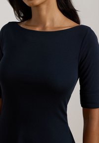 Donna che indossa una maglietta aderente di colore blu scuro con scollatura a barca e maniche a tre quarti su uno sfondo chiaro e semplice.