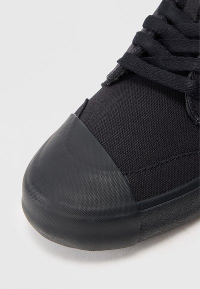 Sneaker nera in tela con punta in gomma, superiore testurizzato e lacci neri piatti. La suola è opaca e leggermente rialzata.