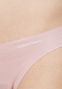 Világos rózsaszín Calvin Klein tanga, amely puha, sima anyagból készült, keskeny derékpánttal és finom, fehér cérnával díszített logóval.