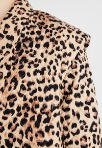 Kviltet stoffjakke med en lys beige bakgrunn og svarte og brune leopardflekkene. Strukturerte skuldre, glatt tekstur og diamantsting.
