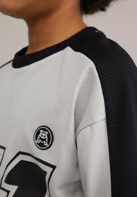Pelēka un melna īsā piedurkne krekls ar apaļo apkakli, ar teksturētu melnu paneli un apļa nozīmīti ar suņa logotipu.