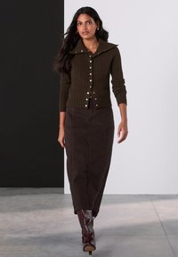 Cardigan oliva a coste con un ampio colletto, chiusura frontale con bottoni, abbinato a una gonna marrone fino alla caviglia e stivali bordeaux al ginocchio.