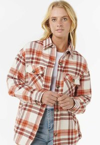Blonde Frau, die ein rot-weiß kariertes Hemd über einem weißen Oberteil und blaue Jeans trägt, steht vor einem schlichten Hintergrund.