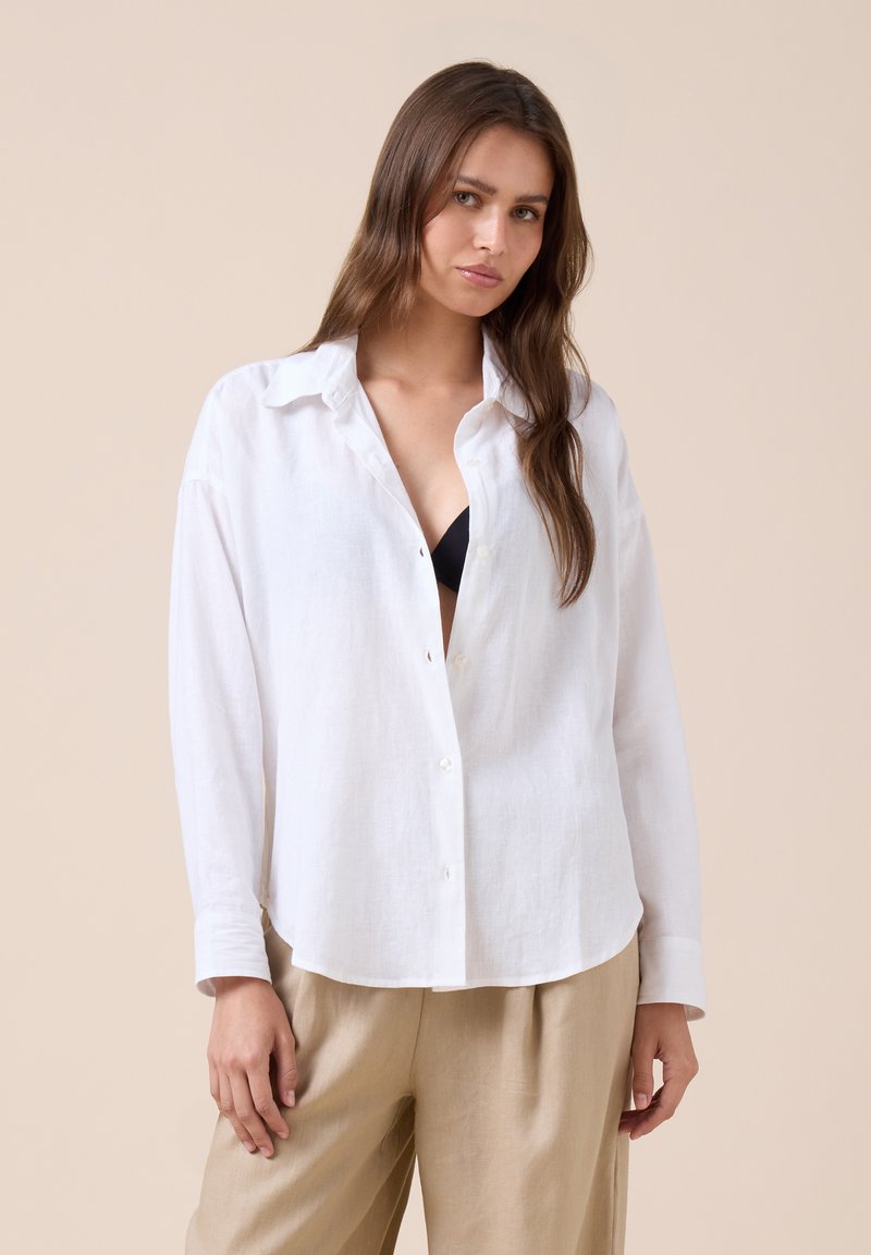 Femme aux longs cheveux bruns porte une chemise blanche boutonnée sur un soutien-gorge noir et un pantalon beige, debout devant un fond beige uni.