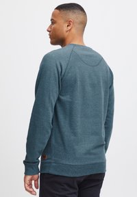 Sudadera en color verde azulado con mangas largas y puños acanalados. Presenta un escote redondeado y un pequeño parche de cuero con el logotipo en la parte inferior izquierda.