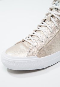Zapatillas altas metálicas doradas con parte superior de cuero texturizado, cordones blancos y suela de goma con un diseño de tracción.