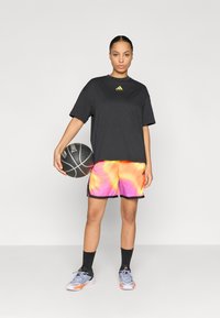 Μαύρο μπλουζάκι με κοντά μανίκια και κίτρινο λογότυπο, πολύχρωμα σορτς tie-dye, μαύρη μπάλα μπάσκετ και γκρι αθλητικά παπούτσια με πορτοκαλί λεπτομέρειες.