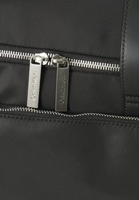 Zaino in nylon nero con due cerniere argentate con etichette Calvin Klein, tasca orizzontale con zip e una finitura opaca liscia.