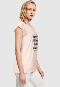 Lyserød bomulds t-shirt med korte ærmer, v-hals, prydet med sort tekst der står "gode ting tager tid" på forsiden. Flad pasform.