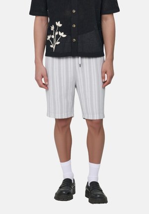 Mann trägt weiß-grau gestreifte Shorts, schwarzes Hemd mit weißen Blumenstickereien, weiße Socken und schwarze Loafers.