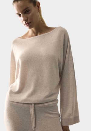Vrouw draagt een beige losse lange mouwen trui en bijpassende broek met trekkoord tegen een effen lichte achtergrond.