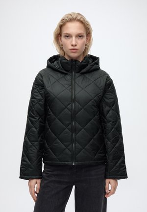 Femme portant une veste matelassée noire à capuche avec fermeture éclair à l'avant et un pantalon noir, debout devant un fond clair uni.