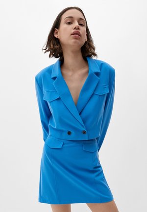 Blaues doppelt-knopfbares Sakko mit großen Revers und zwei vorderen Taschen, kombiniert mit einem passenden Minirock. Glattes Material und figurbetontes Design.