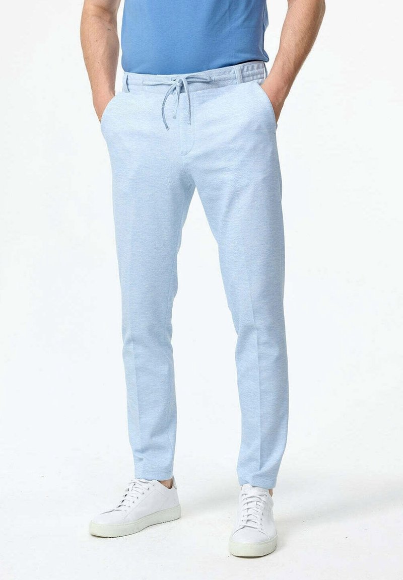 Homme portant un pantalon à cordon bleu clair, une chemise bleue et des baskets blanches, debout les mains dans les poches sur fond blanc.