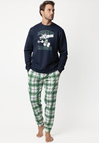 Hombre vestido con sudadera azul marino de Mickey Mouse y pantalones de pijama a cuadros verdes, de pie descalzo con las manos en los bolsillos, mirando a la derecha.