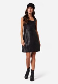 Robe sans manches en cuir noir avec un décolleté carré, corsage ajusté, jupe évasée et coutures minimales. Associée à des bottines noires.