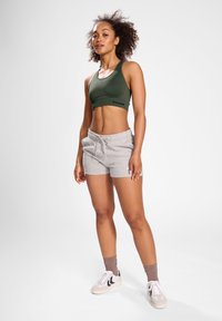 Sportowy stanik w ciemnozielonym materiale, zestawiony z jasnoszarymi szortami. Model nosi białe buty z czarnymi akcentami oraz brązowe skarpetki kostkowe.