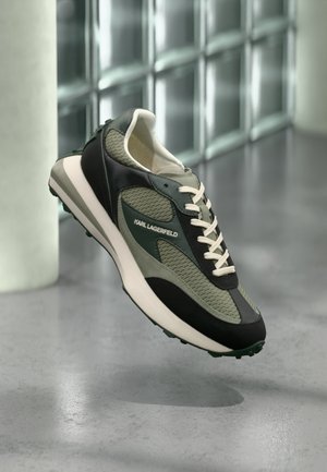 Zapatilla deportiva Karl Lagerfeld en verde y negro con cordones blancos suspendida sobre un suelo gris y con una pared de bloques de vidrio de fondo.