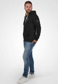Zwarte hoodie met gele accenten, twee voorzakken en witte koordjes. Draagt bij blauwe jeans en witte sneakers.