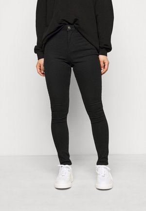 Person iført sorte skinny jeans, sort bluse med lange ærmer og hvide sneakers, der står mod en ensartet lys baggrund.