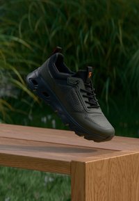 Black athletic shoe with mesh material, textured panels, and a cushioned sole, displayed on a wooden surface against green foliage.