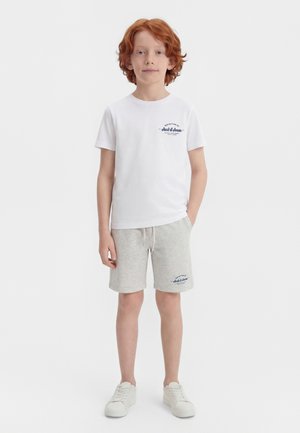 Κόκκινο-μαλλιαρό αγόρι που στέκεται, φοράει λευκό μπλουζάκι και γκρι σορτς με λογότυπα, και λευκά αθλητικά παπούτσια σε λευκό φόντο.