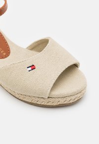 Beige kanva espadril sandaali leveän hihnan, juuttipohjan ja pienen punavalkosiniset kirjontalogon kanssa sivussa.