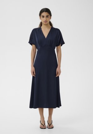 Naine seisab sirgelt, kandes tumesinist lühikeste varrukatega V-kaelusega midi pikkusega kleiti, millele on lisatud mustad kontsadega sandaalid, valgel taustal.