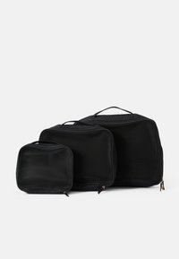Trois organisateurs de voyage en maille noire de tailles variées, dotés de fermetures éclair et de poignées supérieures pour un transport facile. Surface texturée.