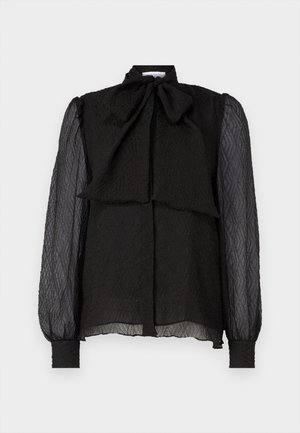 Blouse noire avec un col en nœud structuré, tissu texturé, longues manches transparentes et fermeture à boutons sur le devant. L'ourlet est légèrement arrondi.