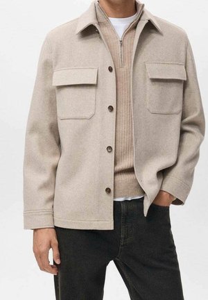 Homme portant une veste beige à boutons avec poches à rabat sur un pull en maille beige zippé et un pantalon noir, main dans la poche.