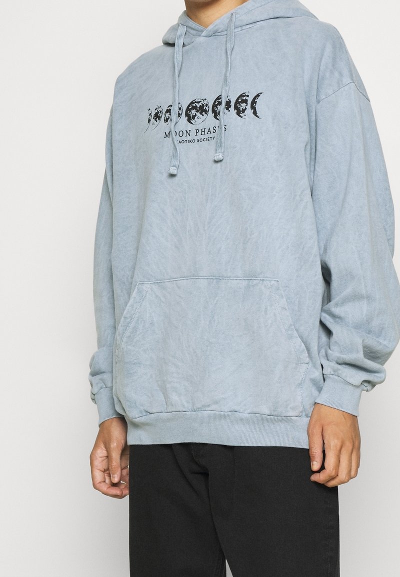 Sweat à capuche bleu clair avec poche kangourou, arborant un graphique noir des phases lunaires sur la poitrine et une capuche à cordon.