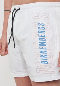 Witte zwembroek gemaakt van lichtgewicht materiaal, met een zwarte trekkoord, zijzakken en gedurfde blauwe en oranje tekst aan de zijkant.