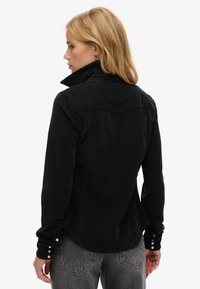 Sort denimskjorte med høj krave, lange ærmer og knaplukninger ved håndleddene. Bagsiden har et rygdesign og en let buet søm.