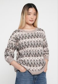 Przesadny sweter w jasnoróżowym kolorze z ciemnoszarymi i kremowymi wzorami w zygzak. Posiada okrągły dekolt i ściągacze. Miękka, teksturowana dzianina.