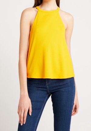 Kobieta w żółtym, prążkowanym, bezrękawnym topie z wiązaniem na szyi, w ciemnoniebieskich dopasowanych dżinsach, stojąca na jednolitym tle.