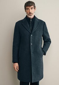 Abrigo de lana gris oscuro con cuello muesca, dos botones, bolsillos laterales y una textura suave, llevado sobre una capa interna negra con cremallera.