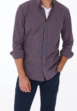 Man met opgerolde mouwen, een geruit overhemd in blauw, rood en wit, en donkerblauwe broek, met handen gedeeltelijk in de zakken.