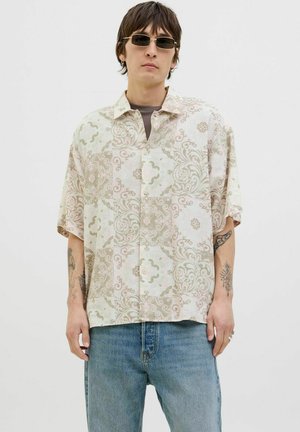 Camisa de manga corta abotonada en tonos claros con un intrincado patrón floral, combinada con jeans de mezclilla y gafas de sol.