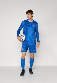 Vīriešu futbolists zilā "Chelsea" formā, turot baltu un melnu futbolbumbu, stāvot pret vienkrāsainu fonu.