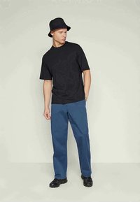 Jovem usando um chapéu preto tipo balde, t-shirt preta, calças azuis e sapatos pretos, de pé contra um fundo claro e liso, olhando para o lado.