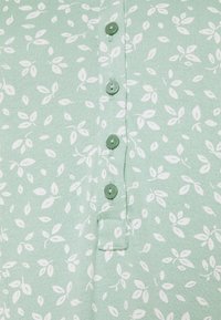 Camicia verde con bottoni, decorata con un motivo foglioso bianco, con sei bottoni verdi e un tessuto morbido e strutturato.