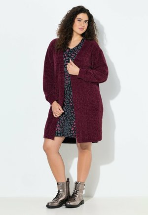 Cardigan texturé bordeaux à ouverture devant, associé à une robe noire à motifs avec des accents roses, et des bottes à lacets métalliques brillantes.